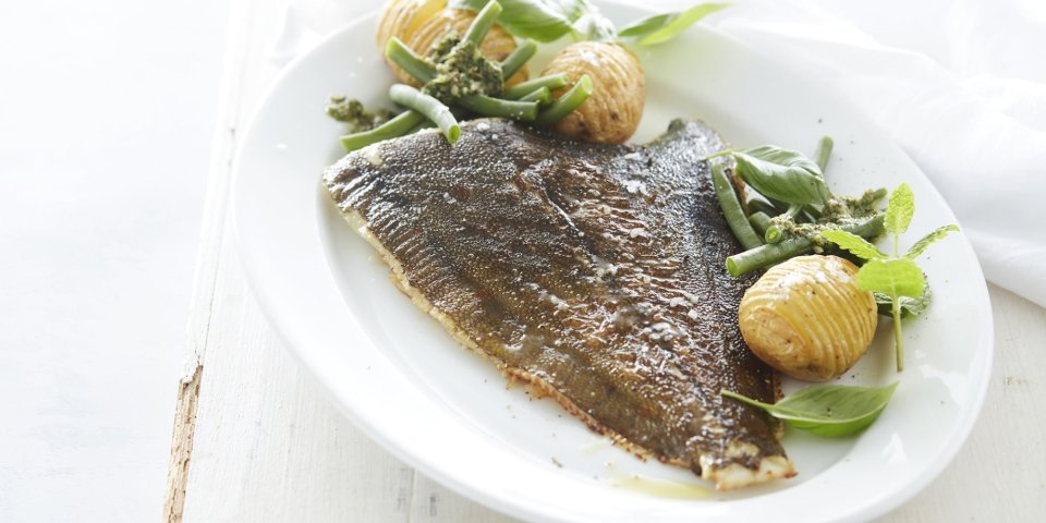 Gebakken pladijs met hasselback aardappelen, boontjes en salsa verde De gebakken pladijs op de graat ligt op een groot wit ovaal bord. Wij serveren hem met boontje langs en enkele hasselback potatoes. Het bord staat op een witte houten tafel en de salsa verde staat apart in een potje boven het bord.
