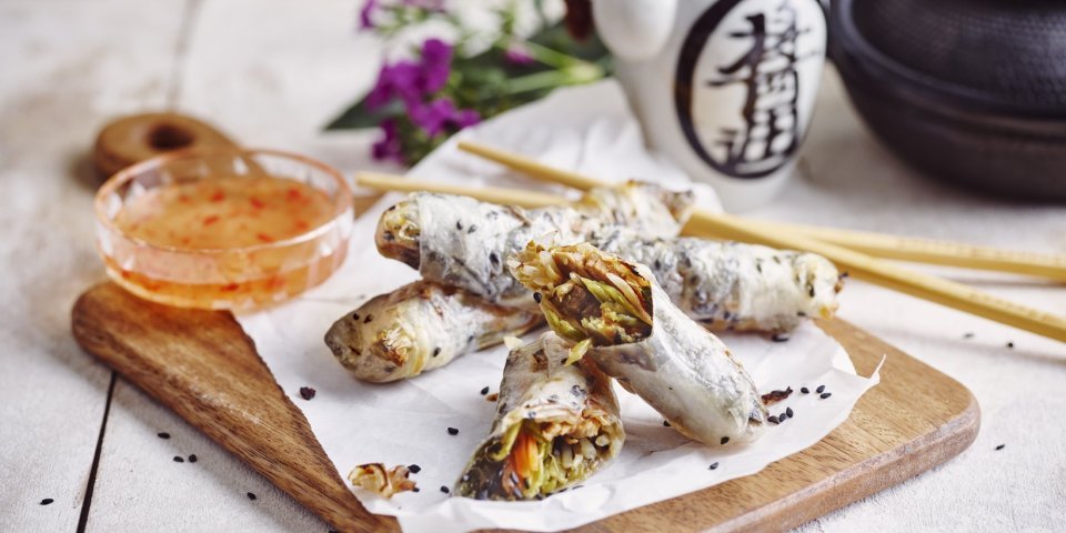 Heerlijke krokante springrolls liggen op een houten snijplank. Ze zijn vergezeld met een zoetpikante dipsaus. In de achtergrond staat nog een theepot en paarse bloemetjes.  