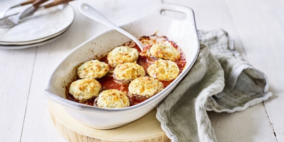 Gegratineerde champignons met blauwe kaas en tomatensaus Witte ovenschotel staat op een houten snijplankje op tafel. De ovenschotel is gevuld met de rode tomatensaus waarin de champignons mooi in geschikt zijn. Achteraan de foto zie je bordjes staan met bestek om aan de slag te gaan.
