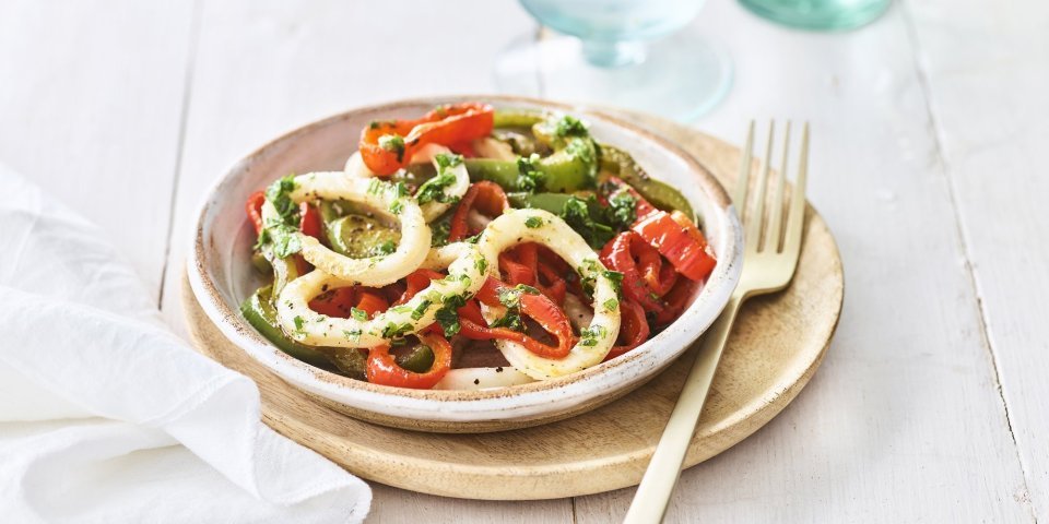 Gegrilde inktvis en paprika met kruidenolie Op tafel staat een keramiek bordje op een houten plankje, het bord is kleurrijk gevuld met groene- en rode paprikaringen, én niet te vergeten de inktvisringen. Een bordje vakantie, als je het mij vraagt.