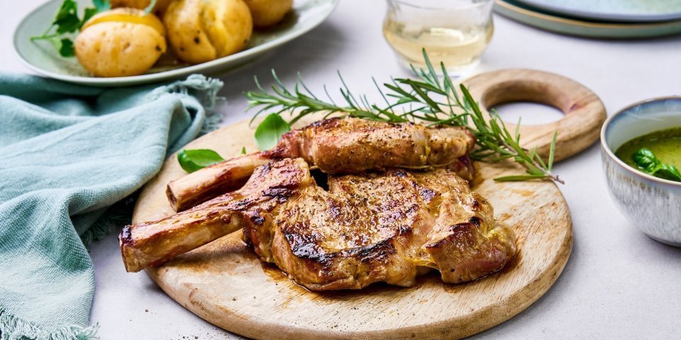 Gegrilde kalfskoteletten met salsa verde op de barbecue Gegrilde koteletten liggen op een houten ronde snijplank te blinken. Er ligt een takje rozemarijn langs, een bordje met de gegrilde krieltjes en een potje met de salsa verde om toevoegen aan het vlees.