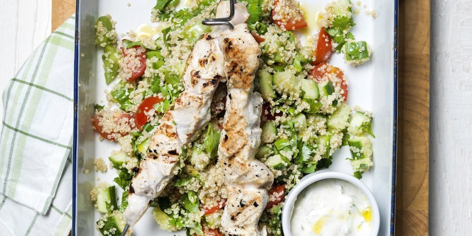 Gegrilde kippenspiesjes in yoghurt met quinoa-salade Grote witte bakplaat staat op tafel met eerst een quinoaslaatje en groentjes gepresenteerd. Hierop liggen de gegrilde brochettes van kip met een dressing van yoghurt.