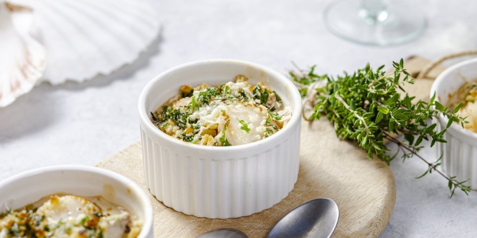 Gegrilde sint-jakobsvruchten met een crumble van crackers, kaas en verse kruiden Gegrilde sint-jakobsvruchten gepresenteerd in kleine witte ovenschaaltjes met een crumble van crackers, kaas en verse kruiden. Ze staan op een houten plankje met kleine lepeltjes en verse tijm naast.