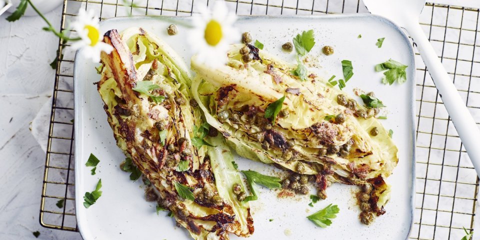 Gegrilde spitskool met ansjovisboter Mooi gegrilde kwarten van een spitskool liggen op een wit dienblad, met hieronder een rooster. Op de spitskool is de ansjovisboter toegevoegd, kappertjes en fijngesnipperde peterselie. Madeliefjes geven de foto een zomerse touch.