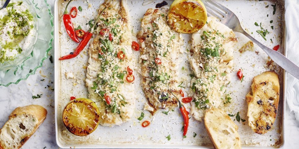 3 stukjes gegrilde vis liggen op een rechthoekige ovenschaal. De gegrilde citroen en chilipepers zorgen voor kleur in dit gerechtje, samen met de groene kruiden in de crumble en dip. 