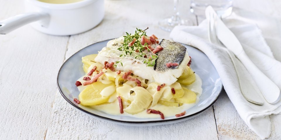 Gepocheerde schelvis in de melk met aardappelschijfjes Schelvis ligt op een torentje aardappelen in een blauw bord. Het bordje staat klaar om aan te vallen op een houten witten tafel met wit bestek langs.