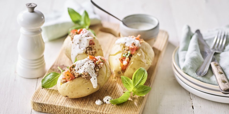 Enkele klaargemaakte en afgewerkte gepofte aardappelen liggen op een houten snijplank. Ze zijn gevuld met gehakt, stukjes tomaat en afgewerkt met een geitenkaasdip. Naast de aardappelen staan enkele bordjes, bestek en servetten om meteen te beginnen. 