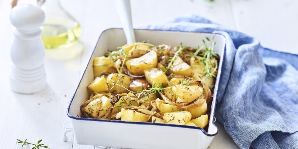 Geroosterde aardappelen met balsamico Een witte ovenschaal met donkerblauw randje staat op een keukenonderlegger op een witte houten tafel. Er staat een witte pepermolen en een glazen flesje met olijfolie langs. De verse tijm zorgt voor extra kleur bij de geroosterde aardappelen, er ligt ook nog een blauwe linnen servette langs de ovenschotel.