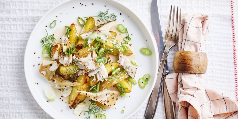 Een heerlijk bordje aardappelsalade staat op tafel, wij serveren hem met stukjes makreel en lente-ui. Om af te werken combineren we hem met een mosterdsausje op basis van yoghurt en mayo. Er ligt bestek langs en roze servette met blauwe lijntjes.