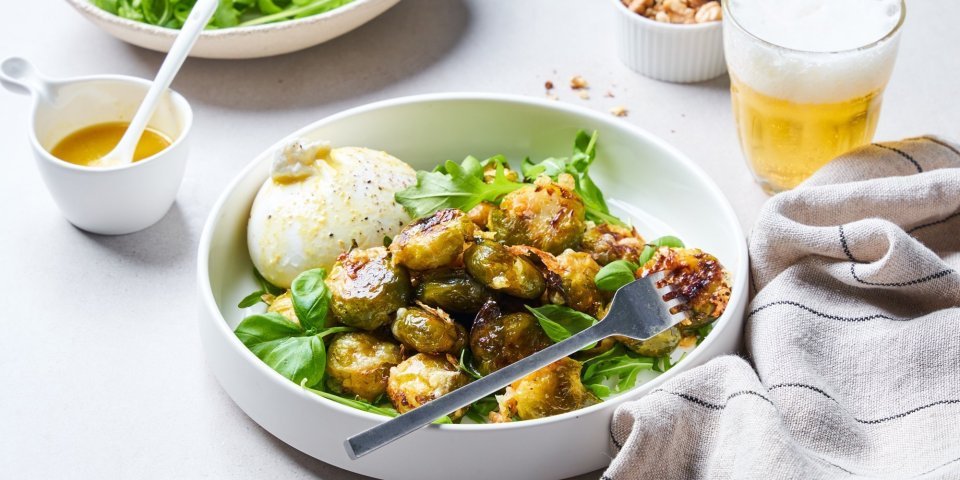 Geroosterde spruitjes met burrata In een diep wit bord liggen de geroosterde spruitjes op een bedje van rucola en basilicum. Hierlangs ligt de burrata. Er is al wat vinaigrette gesprenkeld op de groenten en burrata.