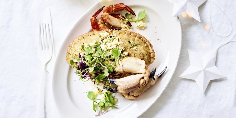 Gevulde Noordzeekrab met krabsalade en vadouvanmayonaise Als het feest is, dan mag het wat meer zijn toch? Gevulde krab met homemade krabsla ligt op een ovaal wit bord met wit bestek lang. Glaasje witte wijn staat bovenaan het bord en er liggen kerstlichtjes en -sterren mee op tafel.