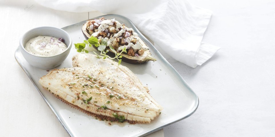 Gevulde aubergine met knolselder, peer en walnoten en schartong Een gebakken schartong ligt op een rechthoekig wit bord. De halve aubergine ligt naast de gebakken vis, hij is gevuld met stukjes knolselder, peer en walnoten. Het gerechtje is afgewerkt met verse gehakte kruiden en een lepeltje zure room.