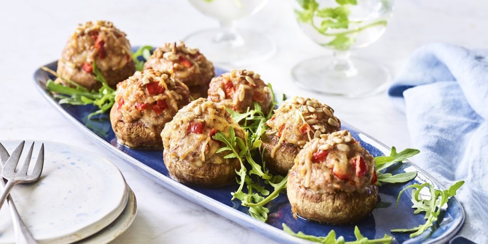 Gevulde champignons met gehakt, paprika en zonnebloempitten Op een blauw schaaltje liggen gevulde champignons. Ze zijn geserveerd met enkele blaadjes rucola en een stappeltje bordjes met mini-vorkjes om het hapje proper te eten.