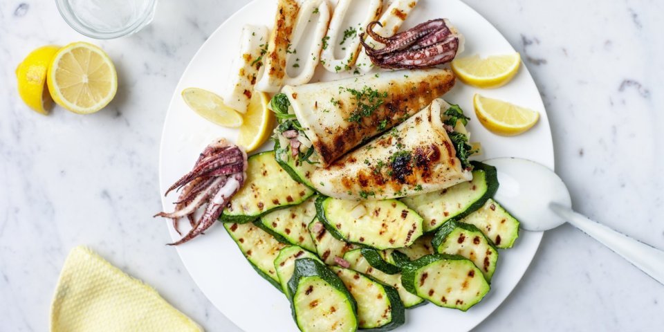 Gevulde inktvis met gegrilde courgette Groot wit bord met gegrilde courgetteschijfjes en gevulde inktvis. Er zijn partjes citroen toegevoegd die zorgen voor het lentegevoel in het beeld samen met de pastelgele servette.