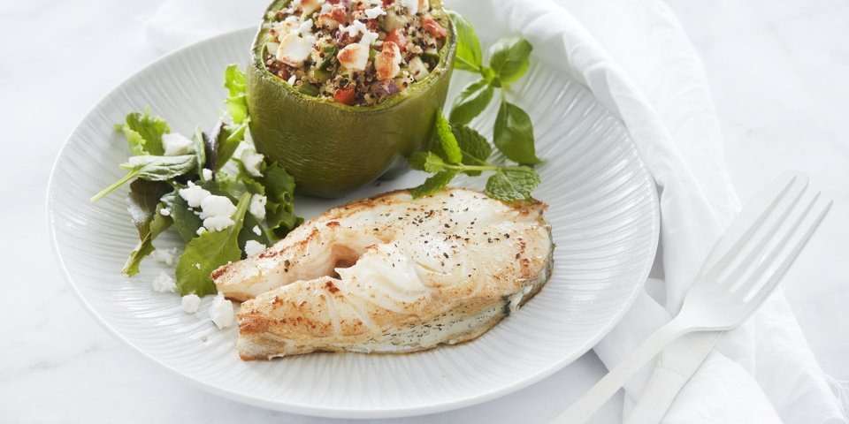 Schelvis met gevulde paprika met quinoa en geitenkaas Een gebakken moot van schelvis ligt op een wit bord. Hij is geserveerd met een gevuld groene paprika. Deze is opgevuld met quinoa, tomaten- en komkommerstukjes en geitenkaasblokjes.