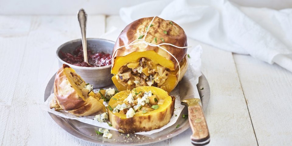 Gevulde pompoen met aardappel, prei en blauwe kaas Een butternut-pompoen ligt op een schaal van inox. Hij is mooi opgevuld met een mengsel van krieltjes, preistukjes en blauweschimmelkaas. Wij combineren hem met veenbessencompote om een zoete smaak toe te voegen.