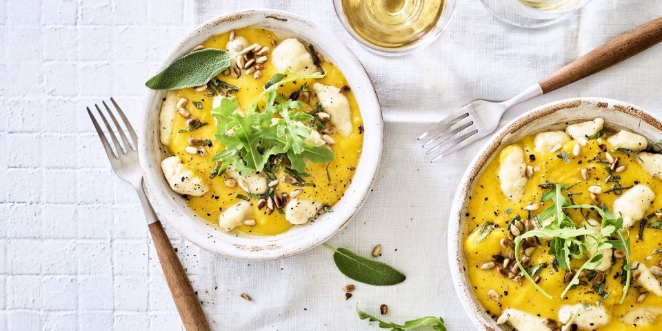 Gnocchi met pompoensaus en salie Gnocchi met pompoensaus en salie staan klaar op een tafel met wit tafelkleed. Bestek met houten afwerking ligt bij het bord, het gerechtje is afgewerkt met verse blaadjes salie en pitten.