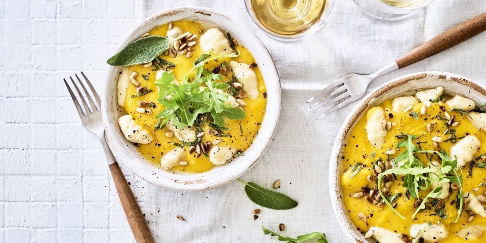 Gnocchi met pompoensaus en salie staan klaar op een tafel met wit tafelkleed. Bestek met houten afwerking ligt bij het bord, het gerechtje is afgewerkt met verse blaadjes salie en pitten. 