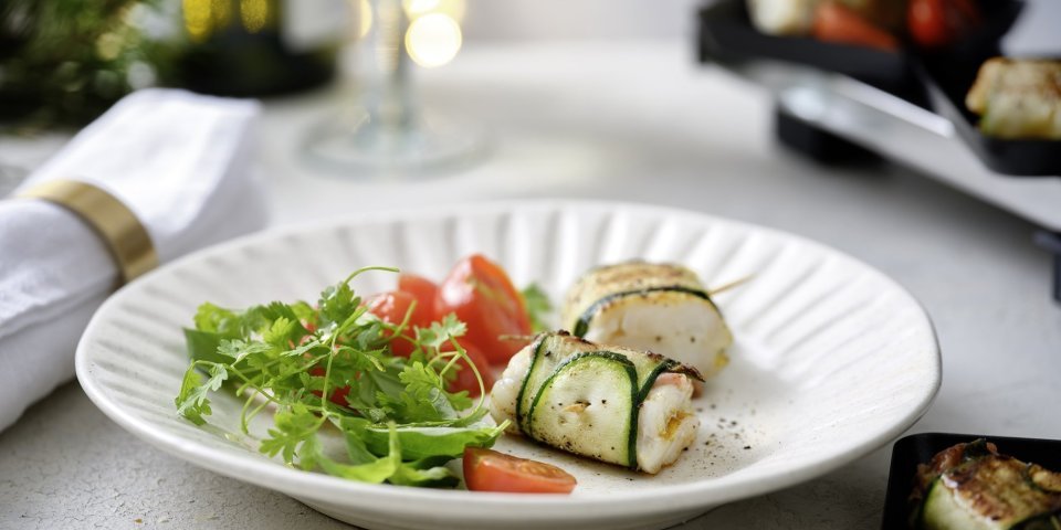 Gourmet van wijtingpakketje met gepofte kerstomaatjes Twee pakjes liggen op een wit bord. Het pakje is gemaakt van een stukje vis verpakt in een sliertje courgette. We serveren dit feestelijk gerechtje met een frisgroene kruidensla en kertomaatjes.