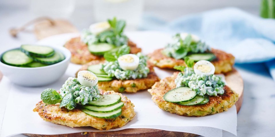 Groentekoekjes van pastinaak, courgette en erwtjes op een houten plank gepresenteerd. Er ligt een licht blauwe linnen servette langs en een waterkaraf met glazen.