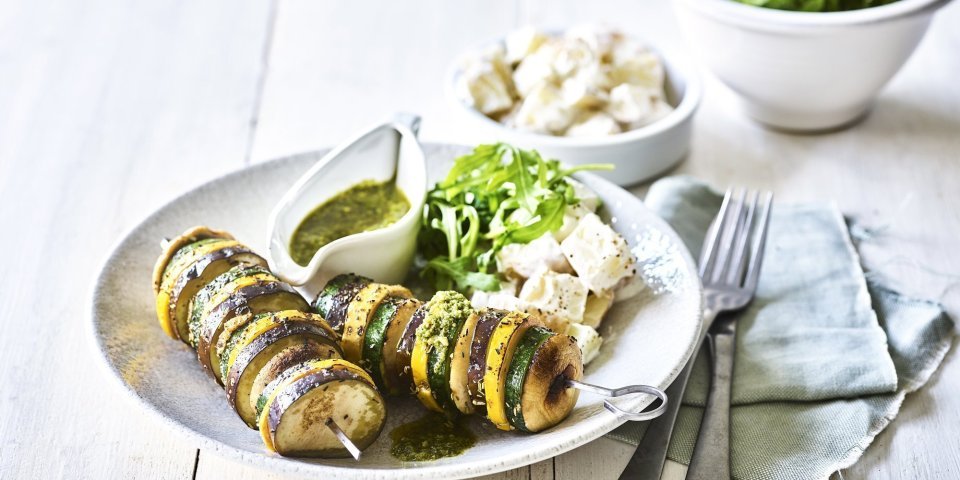 Groentespiesjes met aardappelsalade Op een bleke houten tafel staat een diep bord met hierin een schep aardappelsalade in, twee groentespiesjes met schijfjes van gegrilde aubergine, courgette, champignons en een kannetje met rucolapesto. Verder staat er ook nog een kom met verse groene salade, bestek en een muntkleurige servette.
