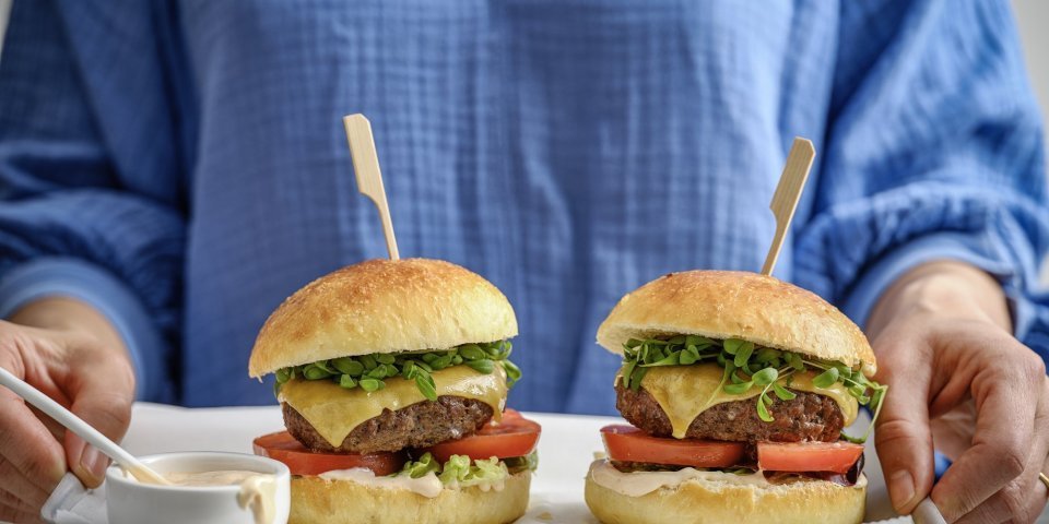 Hamburger Potato buns Een vrouw staat in beeld met haar romp draagt een plateau met hierop twee hamburgers. De hamburgerbroodjes hebben een mooi goudgele kleur, ze werden gemaakt met aardappelpuree in het deeg. Verder zijn de burger belgd met een schijfje vlees, kaas, tomaat, sla en saus natuurlijk.