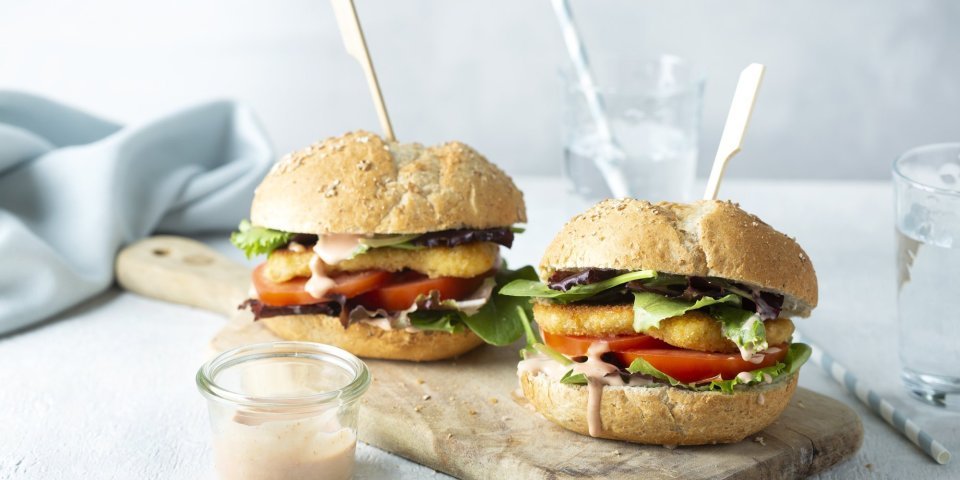 Hamburger met berloumi Een heerlijke hamburger die niemand koud laat. We maakten hem extra crunchy door de kaas te paneren alvorens hem te bakken. De twee hamburgers liggen op een houten snijplank met een prikker middendoor.