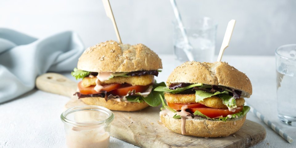 Een heerlijke hamburger die niemand koud laat. We maakten hem extra crunchy door de kaas te paneren alvorens hem te bakken. De twee hamburgers liggen op een houten snijplank met een prikker middendoor. 