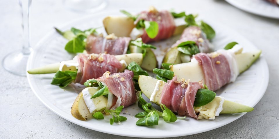 Hapje van peer, Ganda, brie en walnoot Op groot wit bord ligt het hapje geserveerd, de partjes peer met een stukje brie omwikkeld met een sneetje gandaham. Simpel gerechtje, boordevol smaak en kleuren. De groene verse waterkers maakt het volledig af.