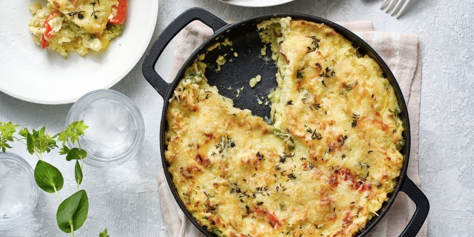 Een taart van aardappelpuree en paprika, staat op het midden van de tafel. Hij zit in een zwarte schotel en heeft een mooi kaaskorstje meegekregen uit de oven. Er is al 1 portie uitgeschept, en er ligt ook bestek op tafel, sla en verse kruiden. 