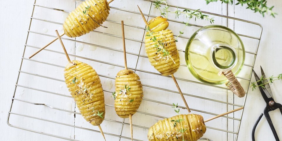 Hasselback potatoes op de barbecue Hasselback potatoes liggen op een bakrooster te blinken. Ze zijn doorprikt met lange houten prikker om makkelijker te kunnen draaien op de BBQ. Er staat een flesje olie, verse tijm en een potje zeezout naast.