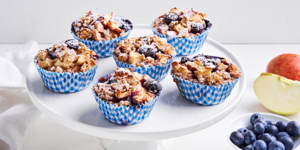 Op een taartenserveerschaal staan 6 muffins in blauw-witte muffinpapiertjes. Er ligt nog een appel langs en blauwe bessen in een potje. 