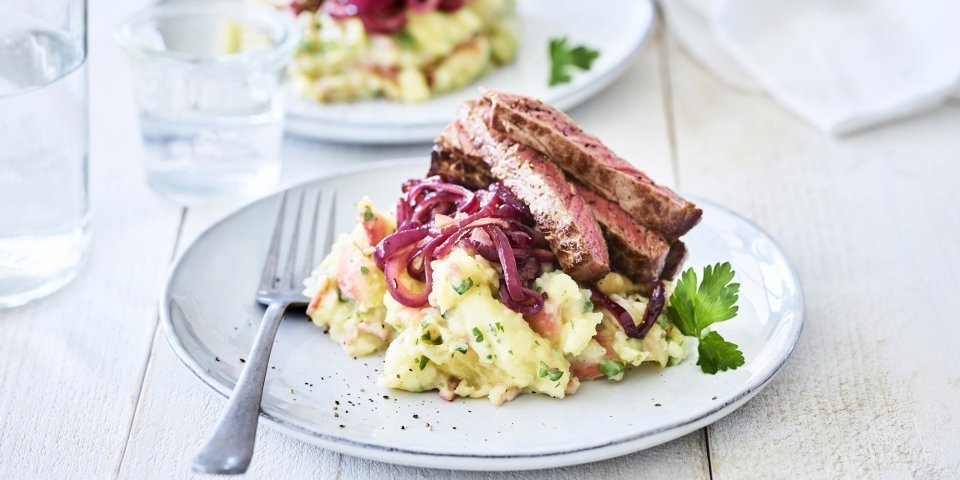 Hete bliksem met biefstuk en rode ui Een lichtblauw bordje is gevuld met een combinatie van puree en appelmoes. Deze puree is afgewerkt met gebakken rode ui en stukjes vlees. Er staan 2 van deze bordjes op tafel met een glas water.