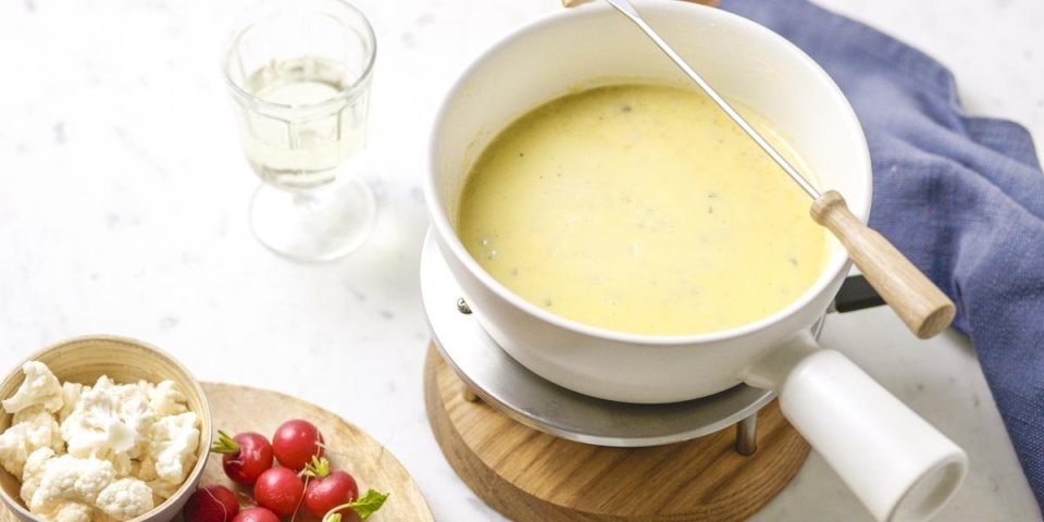 Kassfondue met kazen van bij ons Op tafels staat een fondue-toestelletje met een wit steelpannentje op. Deze is gevuld met de kaasfondue, hierlangs liggen brood, radijsjes, tomaten enz.