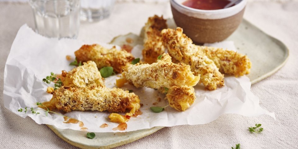 Kaassticks van Brugge Oud kaas met rauwe ham Krokante kaaskroketten liggen op een dienchaaltje, met een dipsausje in een zelfgedraaid keremakiekpotje. Er staan twee glaasjes water naast en de sticks zijn afgewerkt met verse oregano.