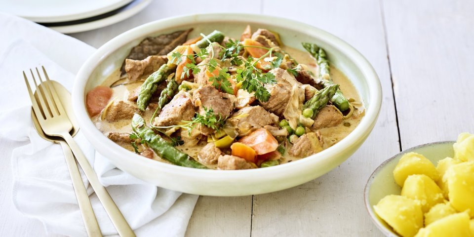 Kalfsfricassee met lentegroenten Deze fricassee straalt een en al lente uit! Hij is klaargemaakt met jonge worteltjes, erwtjes en aspergepunten. Er ligt gouden bestek langs en een portie gekookte aardappelen.