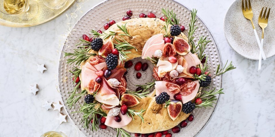 Kerstkrans met charcuterie Een echte Kerstkrans om op te eten. De krans is gemaakt van een Turks brood die belegd werd met verschillende vleesjes, fruit en rozemarijn. Hij staat op de feesttafel en enkele zilveren sterren liggen er langs vergezeld met enkele aperitiefjes.
