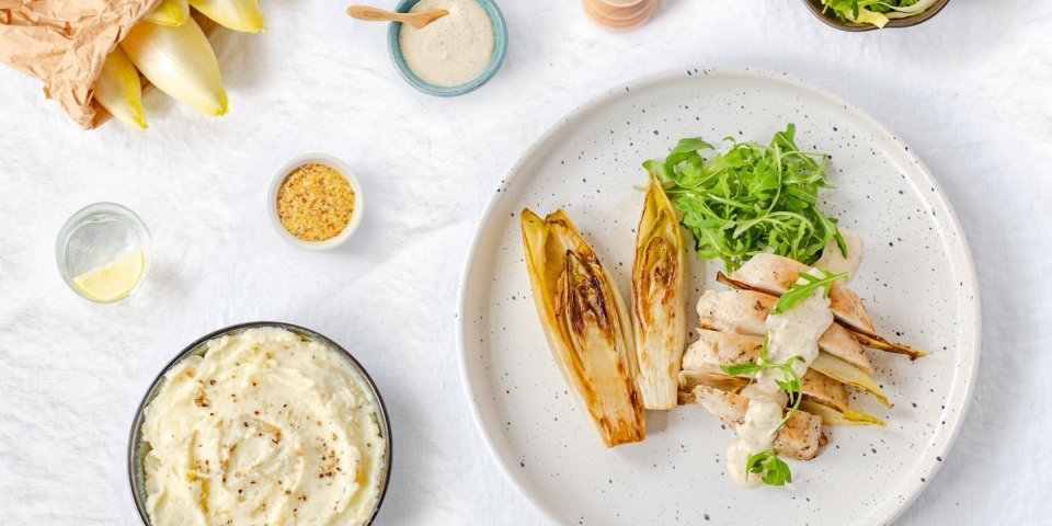 Kipfilet met mosterdsausje en witloof Gestoofd witloof ligt op een wit bord, samen met de kipfilet en mosterdsaus. De groene rucola zorgt voor een frisse kleur in dit gerecht.