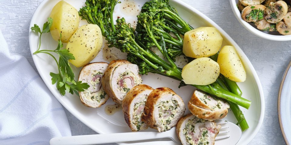 Kiprollade met broccolini en champignons Kipfilet gevuld met een mengsel van roomkaas, mosterd, kruiden en noten. Om af te sluiten voegde we ook nog enkele lapjes spek toe en rolde we hem dicht. Hij ligt aangesneden op een ovaal bord vergezeld van gekookte aardappelen en gekookte broccolini. De gebakken champignons staan in een apart kommetje met een takje peterselie.