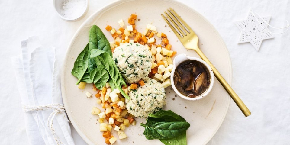 Knoedels met sausje van bospaddenstoelen, gestoofde peterseliewortel en zoete aardappel Knoedels, gemaakt van brooddeeg, liggen bovenop gestoofde blokjes peterseliewortel en zoete aardappel. Deze vegetarische ballen worden opgediend met een sausje van bospaddenstoelen. Het sausje staat mee op het bord in een potje. Goud bestek en kerststerren liggen langs het bord.