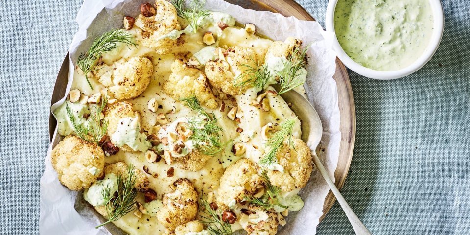 Knolselderpuree met geroosterde bloemkool, groene kruidendressing en noten Puree van knolselder is uitgeschept op een groot lichtbruin bord. Hierop zijn de geroosterde bloemkoolroosjes geserveerd met wat drizzle van de groene kruidendressing, noten en verse dille. De overige kruidendressing is in een witte potje en staat langs het bruine bord, samen met nog wat gehakte noten in een ander potje. Alles staat op een een plaid in een blauwgrijze kleur.