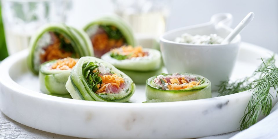 Rolletjes van komkommer gevuld met cottage cheese, wortel, rucola en rosbief. Een heerlijke gezonde en kleurrijke snack op een bordje van marmer. De overige mengeling met de cheese is geserveerd in een dippotje om eventueel een extra toefje te nemen. 