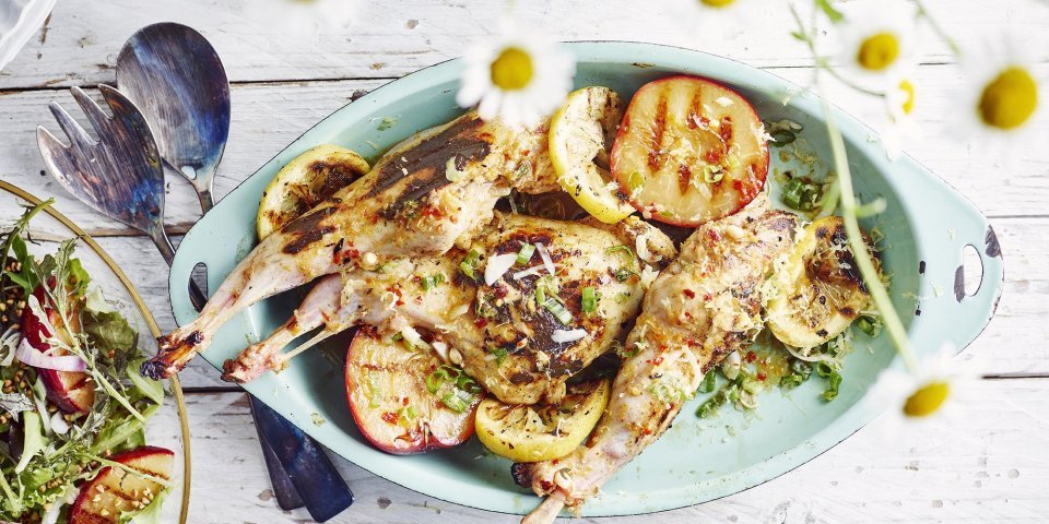 Konijn met gegrilde pruimen op de barbecue Konijn klaargemaakt op de bbq ligt in een lichtblauw bordje samen met de gegrilde halve pruimen. Het slaatje staat links half in beeld op een houten tafel. De foto is opgeleukt met madeliefjes bovenaan onscherp in beeld. Laat de zomer maar beginnen!