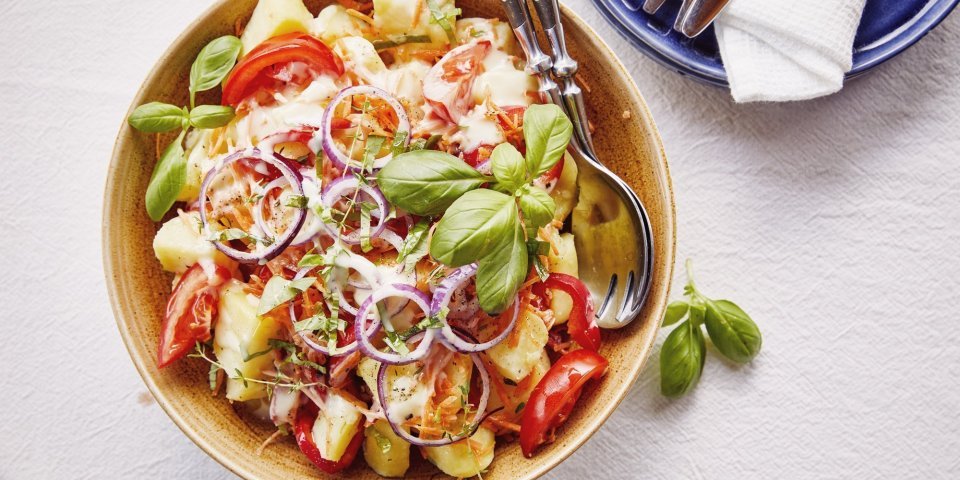 Koude aardappelsalade met tomaat en paprika Een grote serveerschaal staat op de tafel, ze is gevuld met gekookte aardappelstukjes, tomaat, paparika, rode ui en wortel. De dressing is al toegevoegd en verse kruiden zoals basilicum en tijm geven een nog extra kleurtje aan de salade.