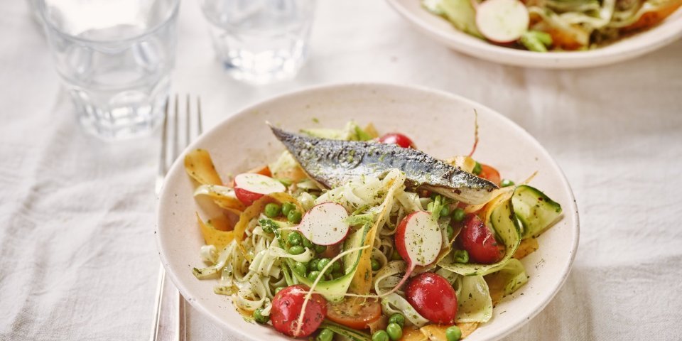 Koude noedelsalade met muntolie, erwtjes en gebakken makreel Een kleurrijk bordje gevuld met groenten, noedels en stukje gebakken vis er bovenop.