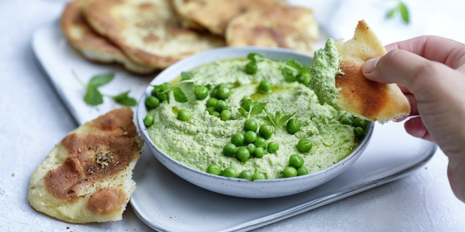 Krokant platbrood met erwtendip Krokant gebakken platbrood ligt op een serveerschaaltje waarbij een dip van erwten en roomkaas in geserveerd. De dip is gemixt en heeft een mooie groene kleur, om af te werken zijn er nog enkele verse erwtjes bovenop gestrooid. Een hand neem alvast een stukje platbrood om te dippen in gezonde groentedip.