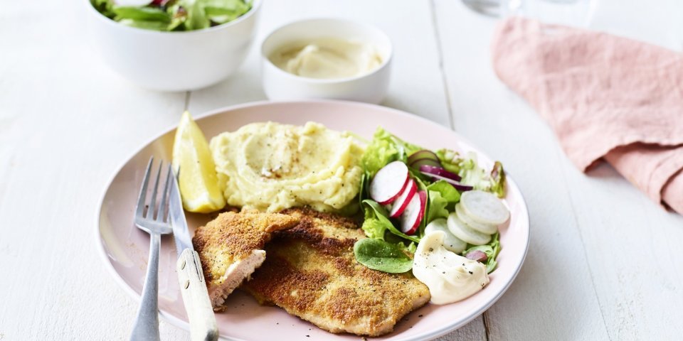 Shnitzel met zelfgemaakte mayonaise Een homemade shnitzel ligt op een pastelroze bord, samen met aardappelpuree, verse sla en zelfgemaakte mayonaise. Er is nog wat extra salade in een wit potje en mayonaise voor extra lekkers op te scheppen.