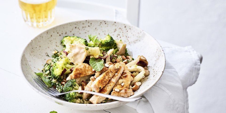 Lauwwarme quinoasalade met groenten, verse kruiden en varkensvlees Een rijkgevulde quinoasalade boordevol verse groenten en kruiden zoals broccoli, paddenstoelen, rode ui, peterselie, koriander en munt. Bovenop de quinoa werd het varkensvlees geplaatst in schijfjes. Er staat een witbier bovenaan het bord, op de tafel.