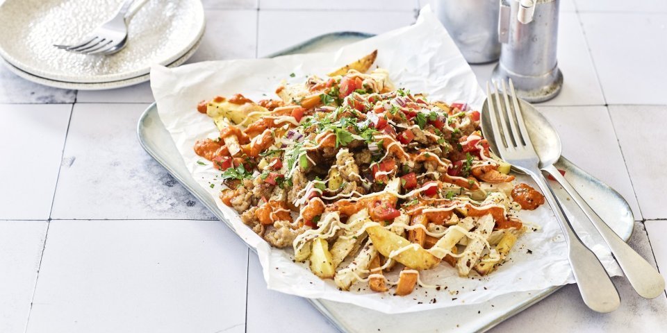 Loaded fries van aardappel, zoete aardappel en knolselder met kip en paprika Op het aanrecht ligt een serveerschaal met een wit bakpapiertje op. Hierop liggen geroosterde aardappel, zoete aardappel en knolselder. Hierop strooien we paprika, gehakt en een sausje. Er staan bordjes langs om op te scheppen en aan te vallen.