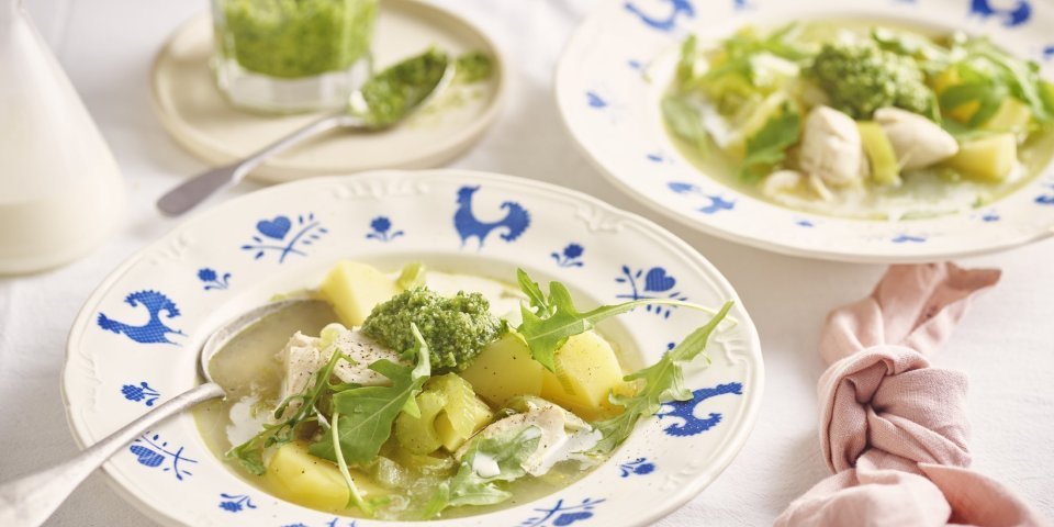 Maaltijdsoep met kip, aardappelen, rucolapesto en room Een wit bord met blauwe kipjes op de rand is gevuld met heerlijke verse kippensoep. Het is een echte maaltijdsoep, met stukjes kip, aardappelen en prei. We werkten de soep af met een scheutje room in het bord en schep rucolapesto. Echt lekker!